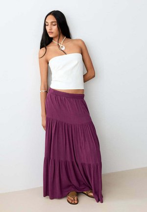 Frau trägt weißes trägerloses Oberteil, langen violetten Stufenrock, Sandalen und Halskette, steht vor einer schlichten hellen Wand.