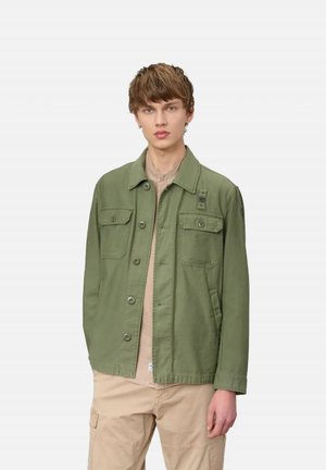 Joven con chaqueta verde oliva abotonada con bolsillos de solapa sobre una camisa beige y pantalones cargo beige, de pie sobre un fondo liso.