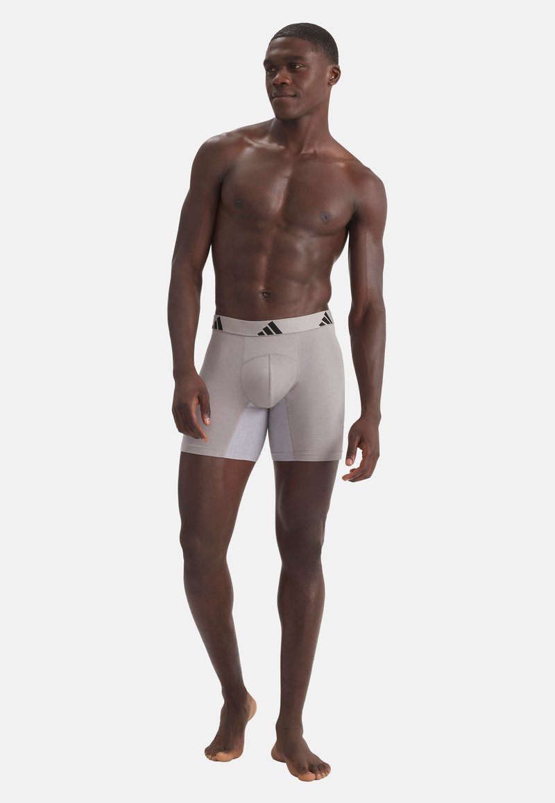Homme athlétique à la peau foncée, debout pieds nus, portant un boxer gris clair Adidas avec le logo sur la ceinture, regardant légèrement sur le côté.