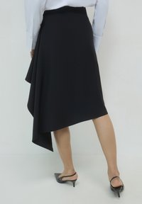 Donna con una gonna midi nera asimmetrica, una camicia bianca a maniche lunghe e scarpe con tacco kitten heel slingback nere, in piedi contro uno sfondo semplice.