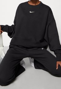 Svart Nike sweatshirt med rund halsringning och en liten vit logotyp på bröstet, i kombination med matchande svarta tapered byxor. Slät struktur.