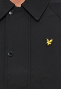 Veste noire avec large col, dotée d'un bouton noir et d'un logo d'aigle brodé jaune sur la poitrine. Texture de tissu lisse.