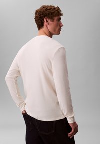 Langærmet, cremefarvet tekstureret skjorte med ribbet mønster. Har rund halsudskæring og tætsiddende manchetter, kombineret med mørke denimjeans.