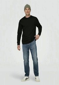 Black long-sleeve shirt, light blue jeans, white sneakers with black accents, and a green knit beanie, standing pose against a plain background.