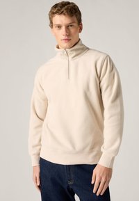 Φούτερ από κρεμ φλίς με όρθιο γιακά, κλείσιμο με ημίκουμπα και μανσέτες με ρίγα. Διαθέτει διακριτική σήμανση στο ύφασμα.