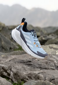 Ανοιχτό μπλε και γκρι αθλητικό παπούτσι Adidas Terrex για τρέξιμο σε μονοπάτι με πορτοκαλί λεπτομέρειες, αιωρούμενο πάνω από βραχώδες έδαφος σε εξωτερικό χώρο.