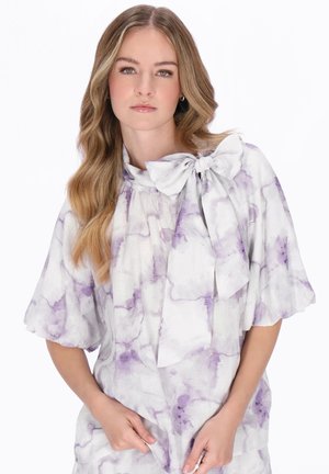 Kobieta z długimi, falującymi włosami, ubrana w białą bluzkę z fioletowymi wzorami akwarelowymi i dużą kokardą na dekolcie, z rękami spoczywającymi w pobliżu bioder.