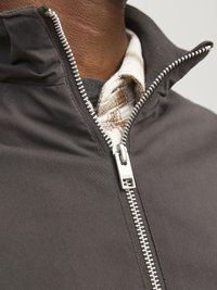 Dark gray jacket with a high collar and a full-length silver zipper. Layered over a plaid shirt with a textured pattern.