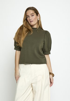 Ženska z dolgimi blond lasmi, oblečena v olivno zeleni top s kratkimi napihnjenimi rokavi in visokim pasom kremnih hlač, stoji pred belim ozadjem.