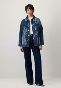 Sieviete, tērpusies tumši zilā divtonīšā iznīcinātā džinsu jakā, baltā krekliņā, tumši zilās augsta jostas kroku biksēs, melnā jostasvietā un brūnās norādītu purngalu kurpēs.