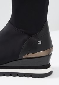 Botte noire à la cheville avec un dessus en tissu lisse, accent métallique sur le talon, semelle en wedge noire texturée et rayures blanches pour le contraste.