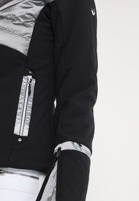 Schwarze und silberne Skijacke mit gepolstertem oberen Bereich, Reißverschlusstaschen und figurbetontem Schnitt. Verfügt über reflektierende Akzente und strukturierten Stoff.