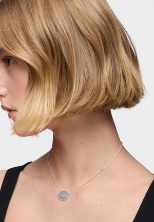 Collana di catena in argento con pendente ovale decorato con accenti arrotondati, indossata da un modello con un taglio di capelli bob mosso.