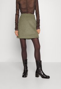 Groene wollen minirok met een rechte vorm. Gepaard met donkerbruine ondoorschijnende panty’s en zwarte glanzende enkellaarzen. Doorzichtige bruine top.