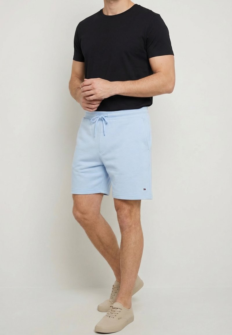 Homme portant un t-shirt noir, un short bleu clair à cordon de serrage et des baskets beiges, debout devant un fond clair uni.