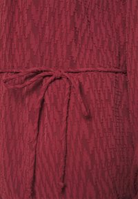 Roter strukturierter Stoff mit erhöhten horizontalen Linien und einem Bindeband in der Taille, das dem Kleidungsstück Struktur und Definition verleiht.