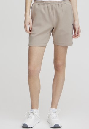 Beige athletic shorts with an elastic waistband, smooth texture, and side pockets. Paired with white sneakers featuring gray accents.