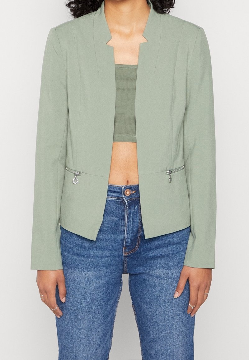 Femme portant un blazer ouvert vert clair par-dessus un top court, avec un jean taille haute bleu, se tenant debout avec les mains détendues le long des côtés.