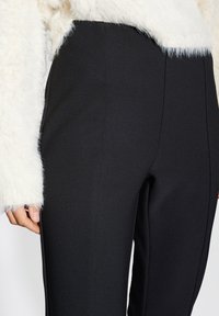 Schwarze, enganliegende Hose aus glattem Stoff, mit einem eleganten Schnitt und sichtbaren Nahtdetails, kombiniert mit einem flauschigen weißen Pullover.