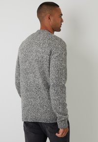 Suéter de punto gris con un patrón texturizado, escote redondo y dobladillo acanalado. Llevado con jeans oscuros, mostrando la vista desde atrás.