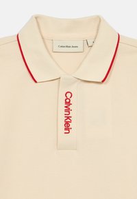 Béžová polo košeľa s červeným akcentom na golieri a vyšitým logom "Calvin Klein". Vyrobená z textúrovanej látky, s klasickým dizajnom a golierom.