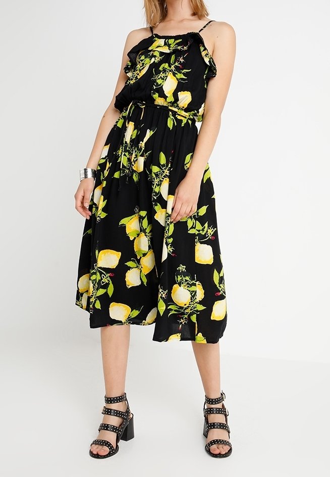Femme portant une robe midi noire sans manches avec un imprimé de citrons jaunes et de feuilles vertes, associée à des sandales à talons blocs cloutés noires et un bracelet argenté.