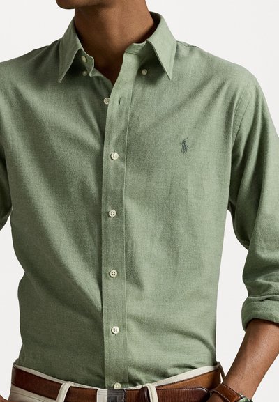 Chemise boutonnée vert clair en tissu texturé, avec un petit logo brodé, des boutons contrastés et un design de col classique.