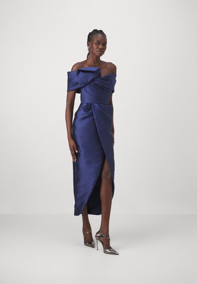 Navy satin off-shoulder dress with a bow detail, side slit, and fitted silhouette, complemented by metallic high-heeled shoes.