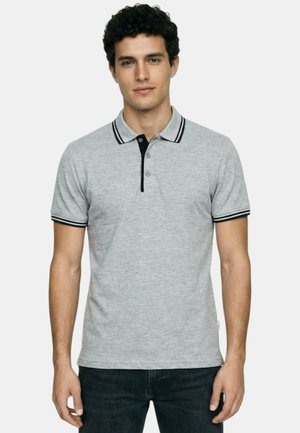 Jeune homme portant un polo manches courtes gris clair avec des bordures noires au col et aux poignets, debout devant un fond blanc.