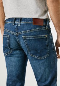 Donkerblauwe denim jeans met een gestructureerde afwerking, contrasterende gele stiksels en een bruine leren patch op de tailleband.
