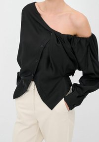 Blusa nera in satin con design off-shoulder, chiusura a bottoni e maniche lunghe e puffate. Abbinata a pantaloni beige.