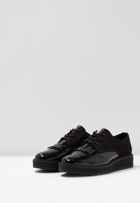 Chaussures noires avec un avant en cuir verni brillant et une tige en daim. Elles sont dotées d'une fermeture à lacets et de franges décoratives. Semelle en caoutchouc épaisse.