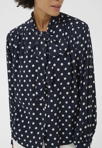 Blouse bleu marine à pois avec un nœud noué au col, en tissu doux, manches longues et détails de boutons au niveau des poignets.