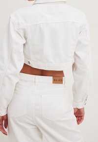 Personne portant une veste en denim blanc raccourcie et un jean blanc taille haute avec une étiquette brune NA-KD sur la taille, vue de dos.