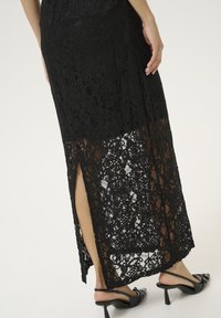 Jupe maxi en dentelle noire avec un ourlet transparent et une fente sur le côté. Texture lisse avec des motifs floraux. Associe avec des sandales à talons noires.
