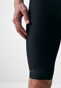 Shorts de sport noirs avec une texture lisse, présentant un design ajusté et un logo discret près de l'ourlet. Main reposant sur le tissu.