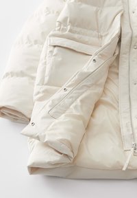Veste à quiltée et rembourrée crème avec une texture lisse et brillante, dotée d'une fermeture éclair à l'avant et de deux poches latérales avec des fermetures à pression.