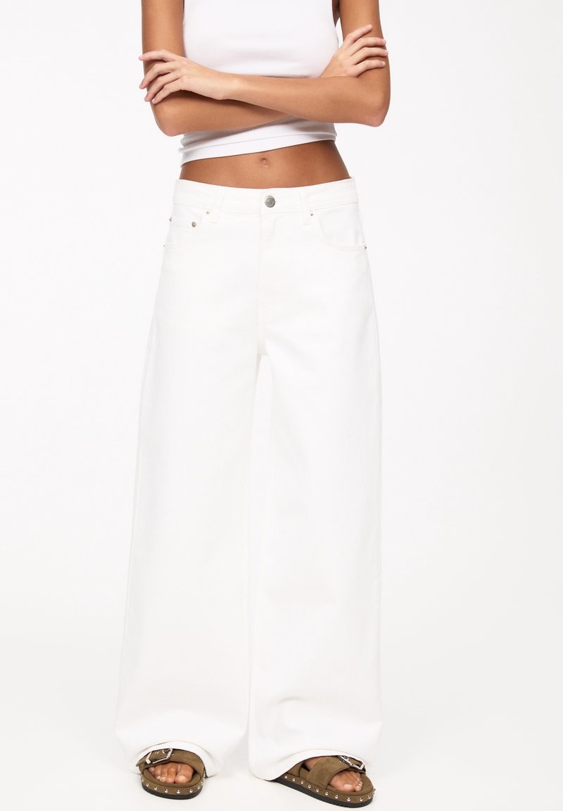Taille fine avec les bras croisés, portant un pantalon blanc taille haute à jambes larges et des sandales marron cloutées sur un fond blanc uni.
