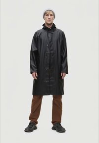 Cappotto impermeabile nero con bottoni a pressione, cappuccio e finitura testurizzata, abbinato a pantaloni marroni e scarpe sportive nere. Sfondo neutro.