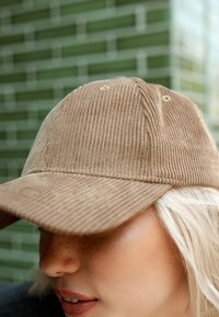 Casquette de baseball en velours côtelé marron avec une visière courbée, comportant des rayures verticales subtiles et des coutures beiges, sur fond de carreaux verts.