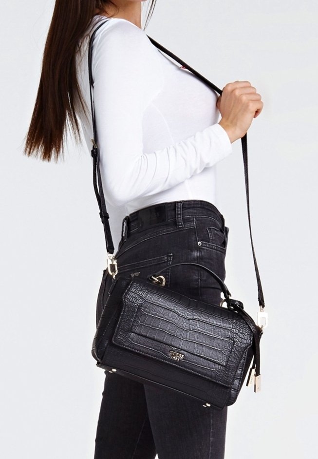 Femme portant un haut blanc à manches longues et un jean noir porte un sac à main noir texturé Guess avec bandoulière.