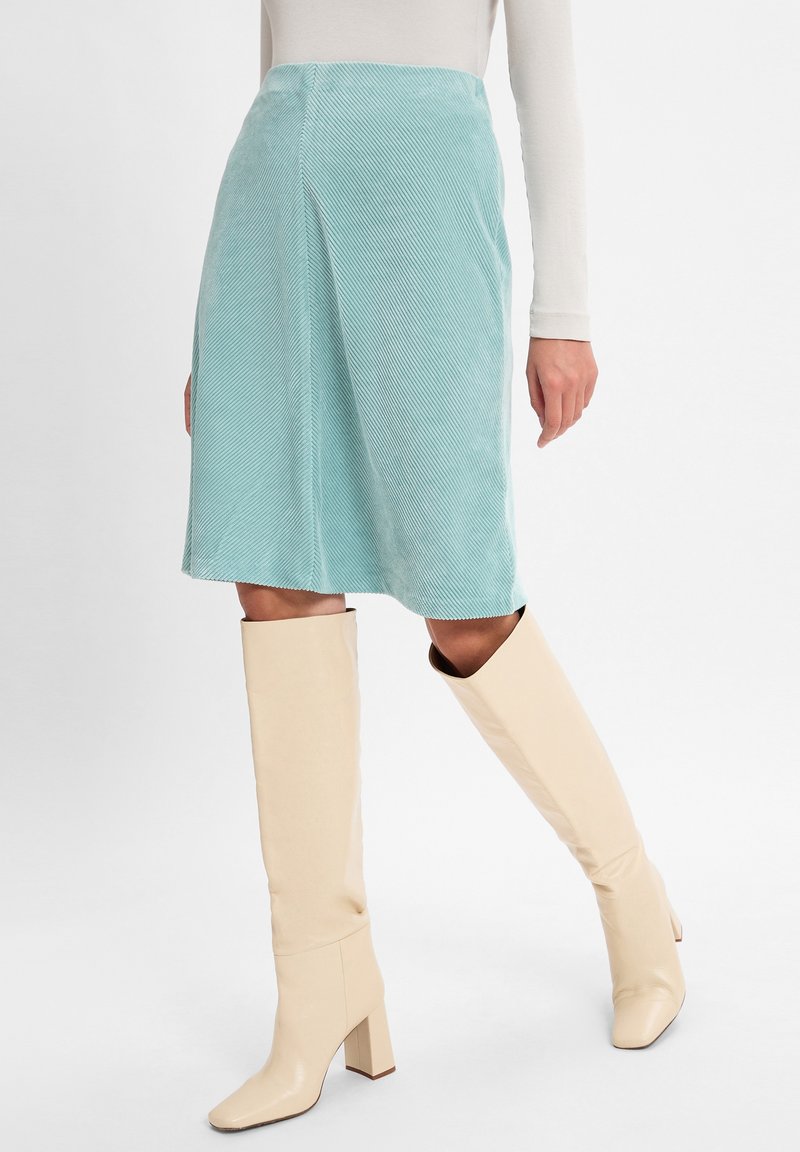 Een teal kleurige corduroy rok met een rechte snit, tot op de knie, met verticale ribbels, gecombineerd met knielange beige blokhaklaarzen.