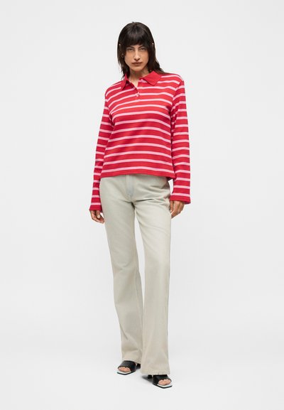 Γυναίκα με μακρυμάνικο πόλο κόκκινο και λευκό ριγέ, μπεζ παντελόνι με φαρδιά μπάτζια και μαύρα ανοιχτά πέδιλα με τακούνι, πόζα σε λευκό φόντο.