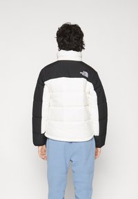 Padded bunda v čiernej a bielej farbe s vysokým golierom. Obsahuje prešívaný dizajn a logo na chrbte. Nosí sa s svetlomodrými nohavicami.