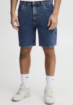 Mørkeblå denimshorts med avslappet passform, med knapp- og glidelåslukking, beltehemper og lommer. Brukt med hvite sokker og joggesko.