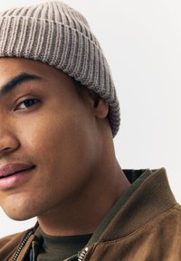 Berretto beige a coste indossato da una persona. Giacca bomber marrone e camicia verde visibili. Primo piano mostra la texture della pelle e i tratti del viso.