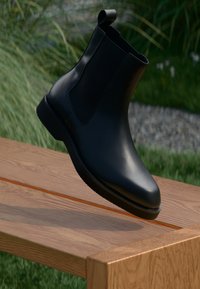 Mustad mustad ankle saapad sileda pinnaga, elastsete külgpaneelidega ja sangaauguga varrel, asetatud puidust pinnale.