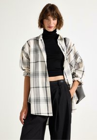 Kvinna klädd i en svart polotröja, stor svart och vit rutiga skjorta, svarta vida byxor och bär en svart väska.