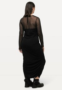 Langes schwarzes Kleid mit durchsichtigen langen Ärmeln, tailliertem Oberteil, gerafften seitlichen Details und abgerundetem Saum. Getragen mit schwarzen Stiefeletten.