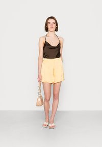 Top halter de cetim castanho combinado com calças de cetim amarelas. A modelo segura uma pequena mala bege com alça de corrente e usa sandálias brancas rasas.